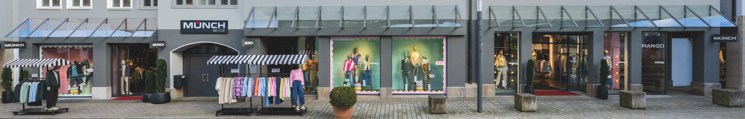 Die Ladenfront von Münch Mode in Künzelsau über die ganze Breite: Schaufenster unter Glasvordächern, Kleiderständer auf dem Gehweg, die Eingänge zu Münch Mode und zum Mango-Shop.